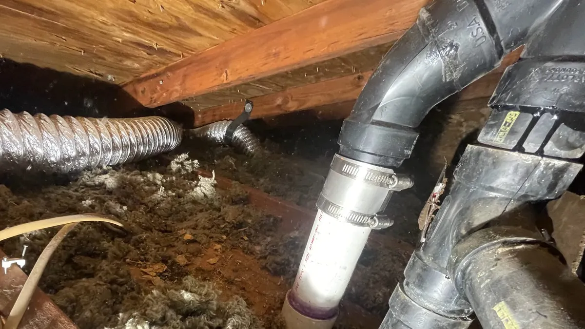 Large vent ducts and white drain piping cross beneath exposed floor framing during a plumbing service call in Maricopa, AZ