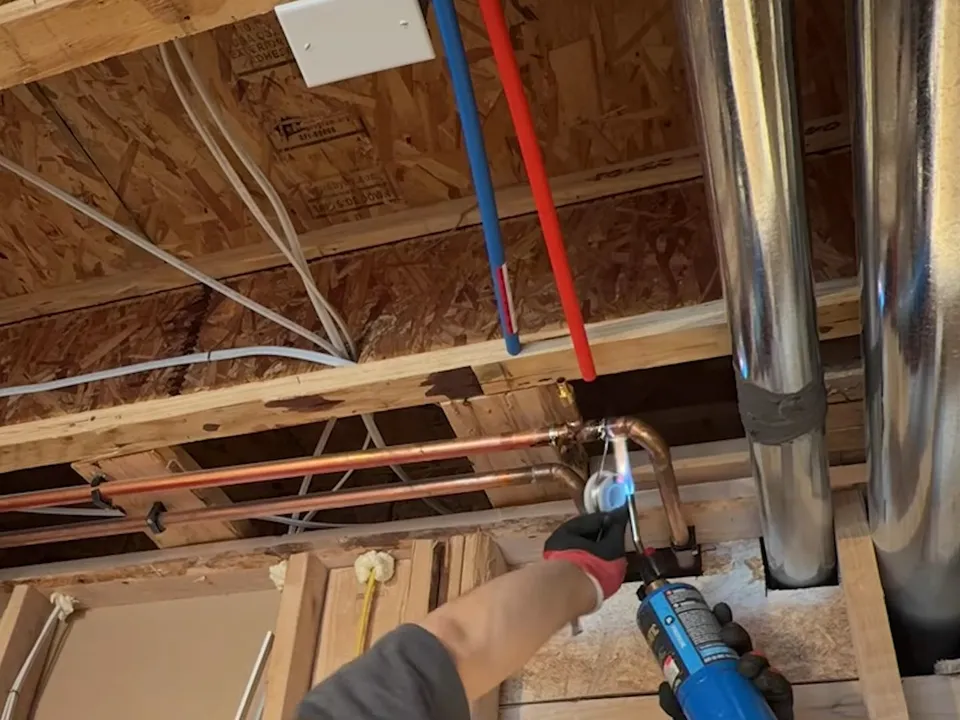 Red and blue supply lines rise toward shiny metal vent piping in an unfinished ceiling during a plumbing service call in Maricopa, AZ