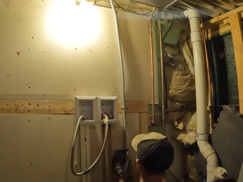 plumber works beside exposed laundry drain and supply piping inside an open wall during a plumbing service call in Maricopa, AZ